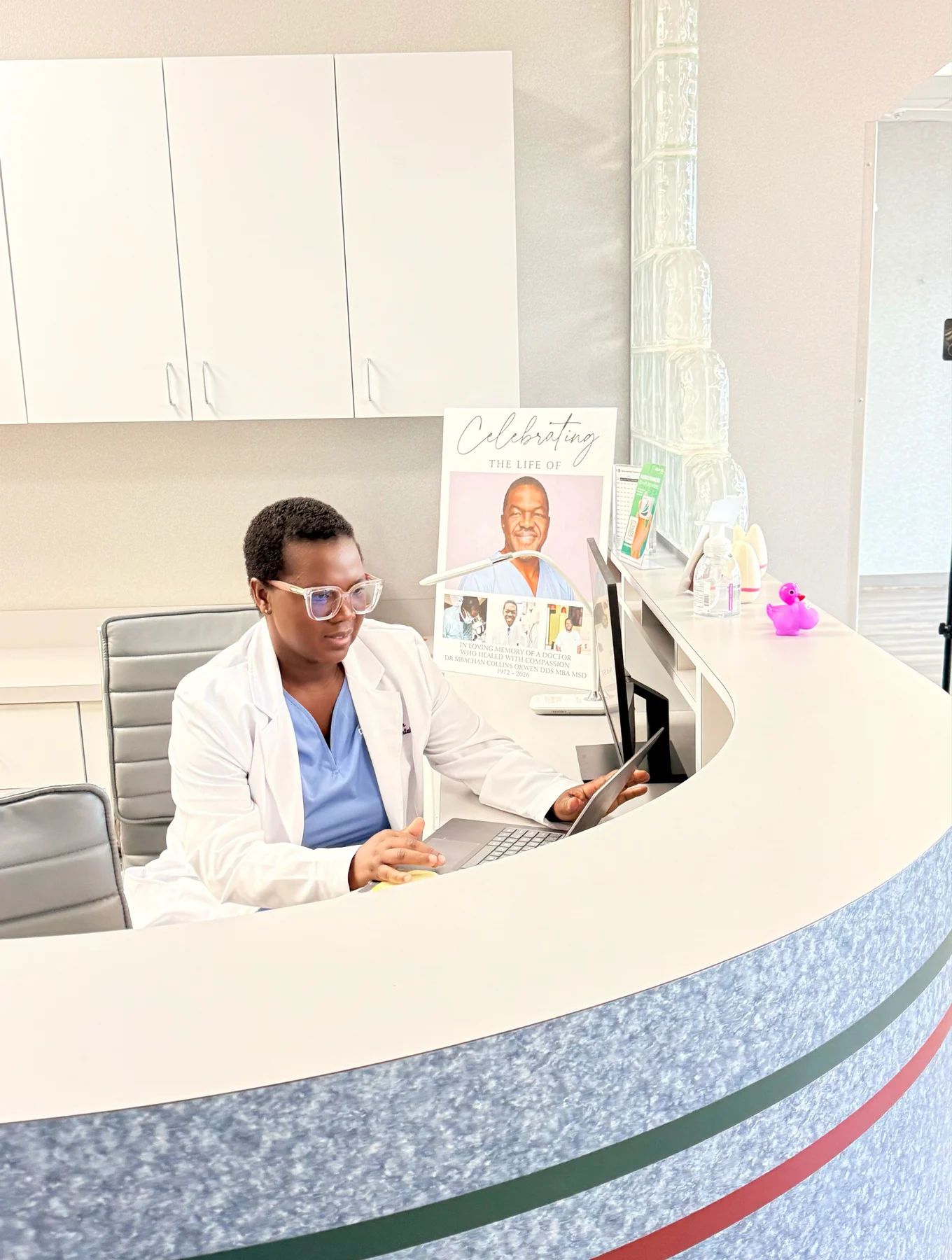 Dr. Fri Vekuh at the reception desk with Dr. Okwen’s memorial portrait behind her